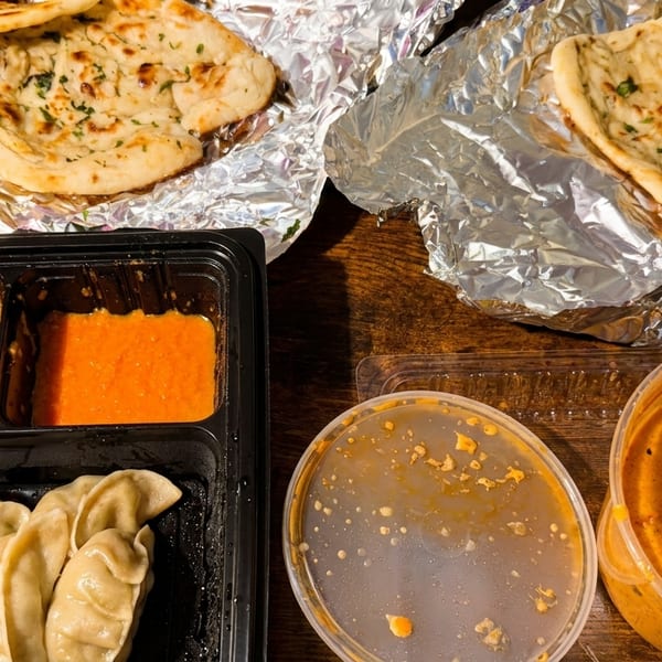 Indian takeout meal with naan bread, momos, and various orange sauces in containers on a wooden table.