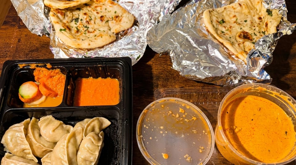 Indian takeout meal with naan bread, momos, and various orange sauces in containers on a wooden table.