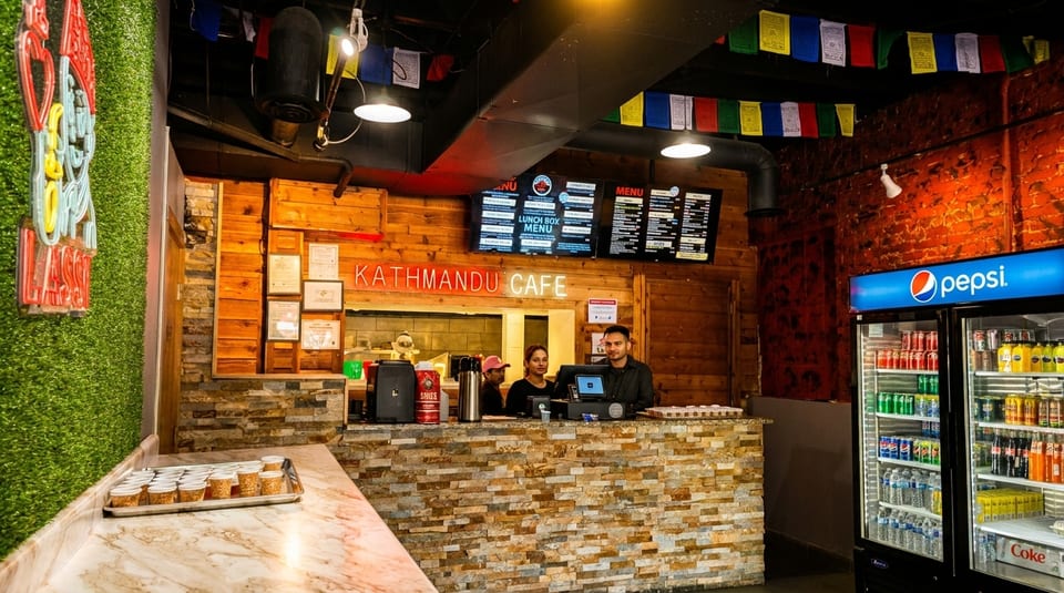 Two staff members stand behind the counter at Kathmandu Cafe, with menus displayed above.