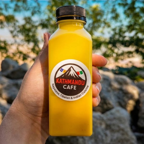 Hand holding a Kathmandu Cafe drink bottle with a beach and rocks in the background.