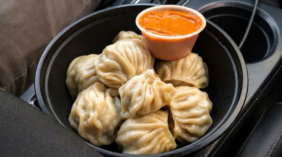 Steamed momos in a black bowl with a side of orange dipping sauce, in a car's cup holder.